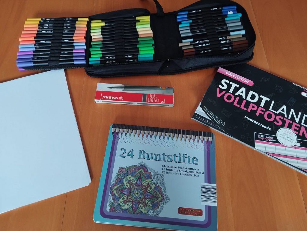 oben ein offen liegendes Stiftemäppchen mit Filzstifetn unten Paket Buntstifte rechts Spielblock "Stadt-Land-Vollpfosten"