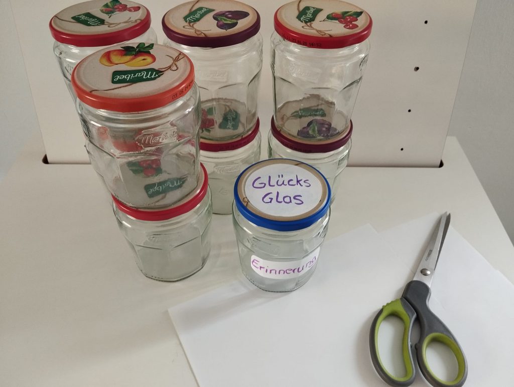links oben leere Marmeladengläser mit dem Aufkleber Glücksglas, unten eine Schere und Papier