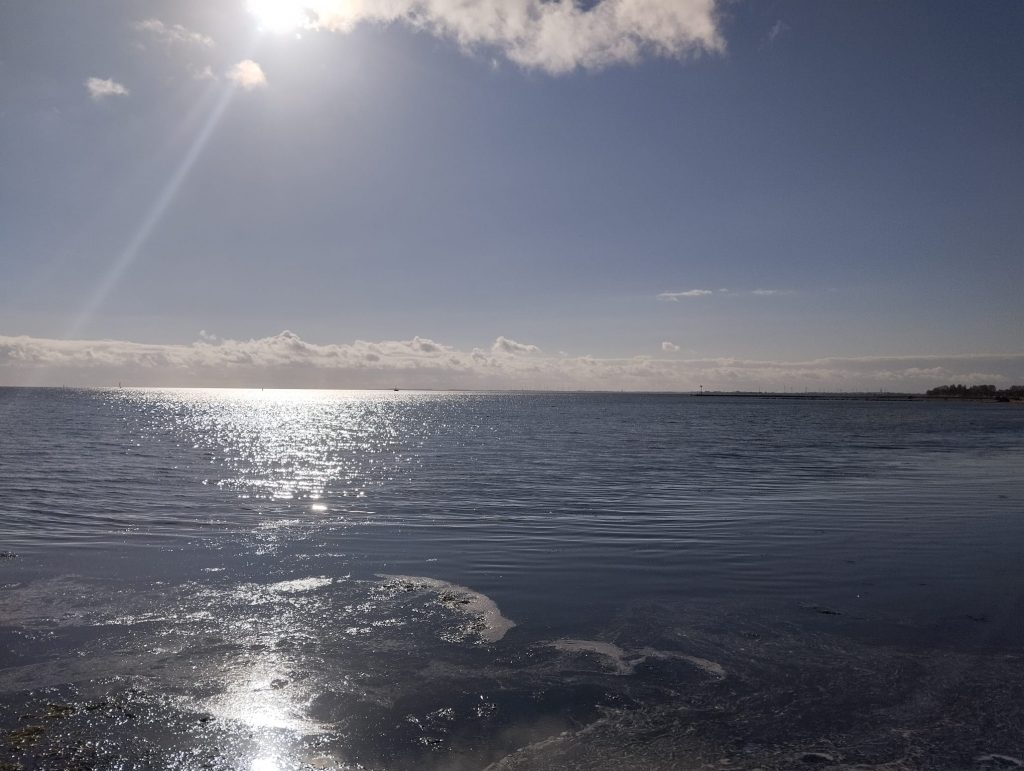 vorne Meer in dem sich die Sonne spiegelt, blauer Himmel mit wenigen Wolken am Horizont