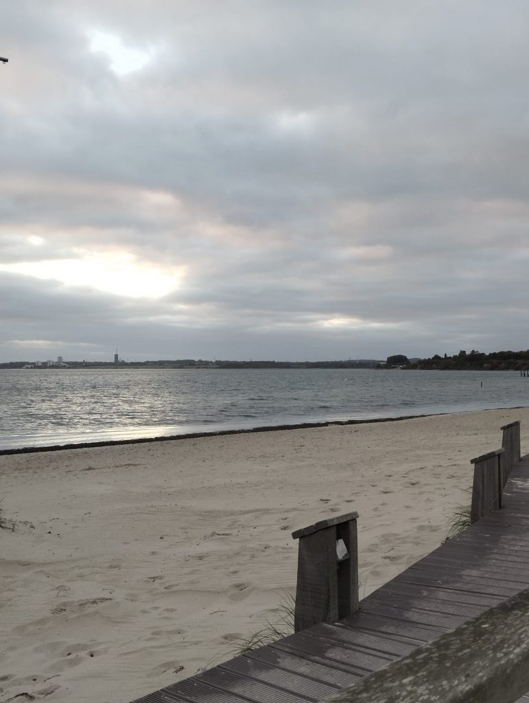 rechts unten Holzsteg links Strand dahinter Meer, bewölkter Himmel durch den die Sonne kommt