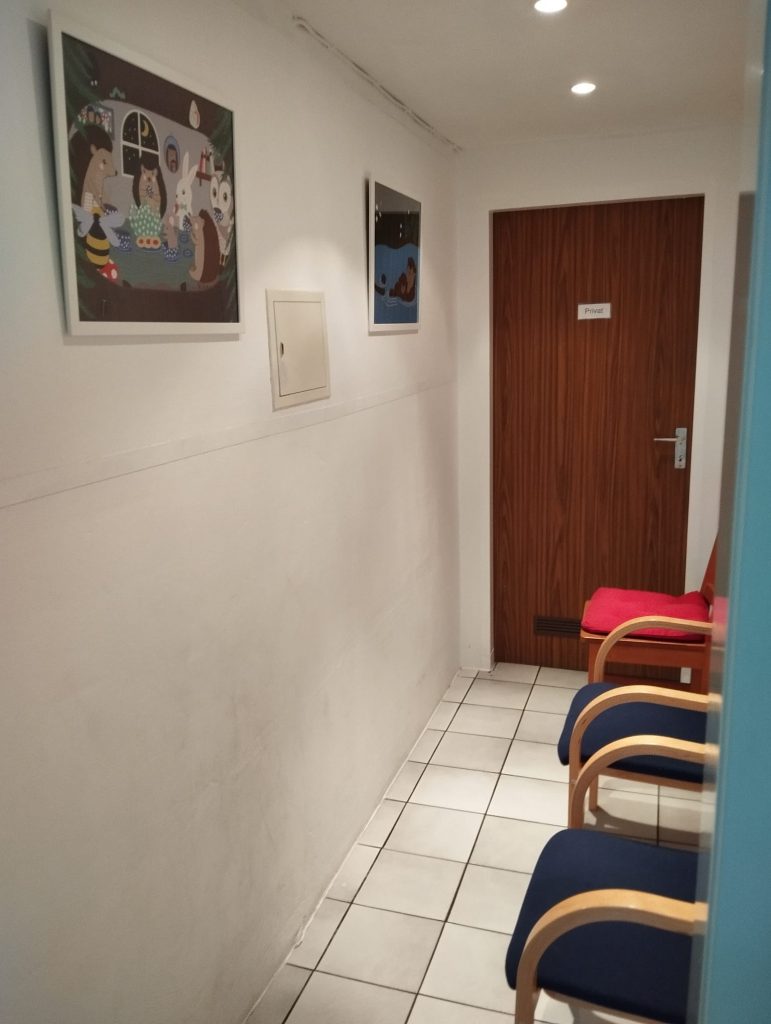 kleiner , schmaler Flur, rechts drei Stühle, hinten eine Tür links zwei bunte Bilder