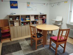 hinten ein Regal darüber Bilder rechts ein Stehtisch in der Mitte ein runder Tisch mit drei Stühlen