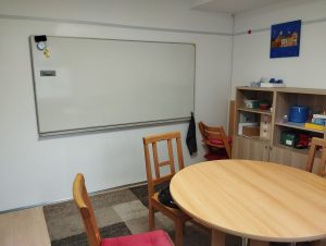 an der Wand ein großes Whiteboard rechts ein Regal vorne ein Tisch mit Stühlen